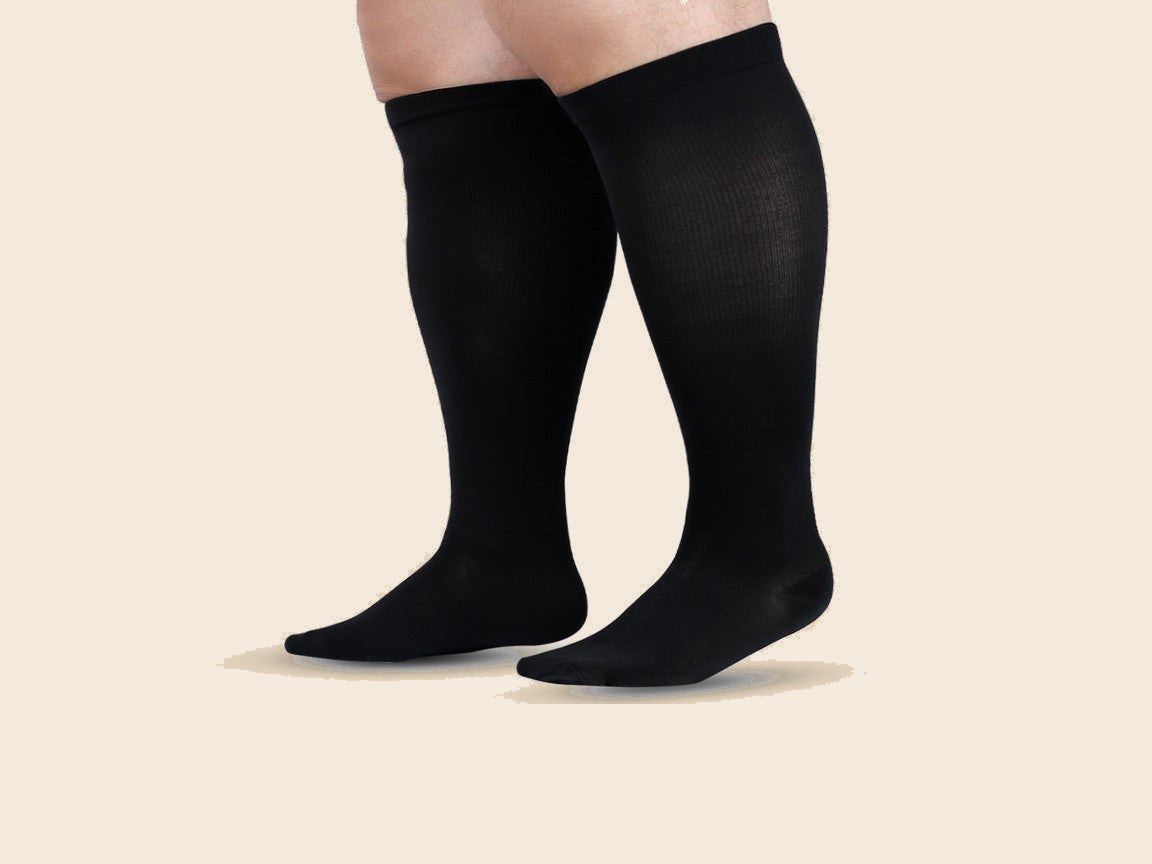 Wide calf compression socks in black, worn on a pair of fuller calves, on a beige studio background