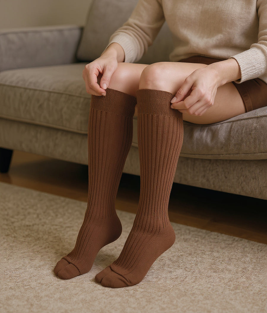 Both legs in brick red knee-high CushyCalf socks, pulling the cuff up while sitting on a sofa at home